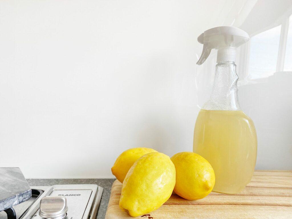 An electric-clear spray bottle filled with lemon liquid sits on a wooden board beside three lemons.