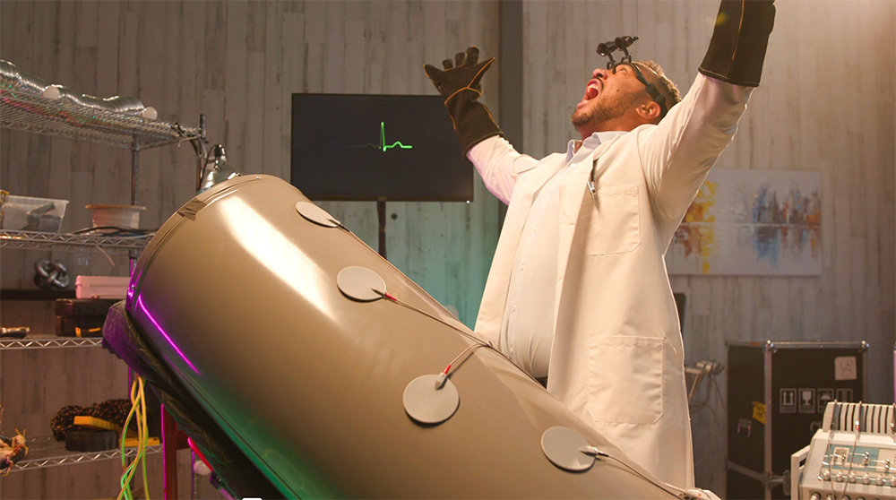 A scientist in a lab coat and gloves raises his arms beside a large cylindrical machine with electrodes, looking up; lab gear and monitor behind.