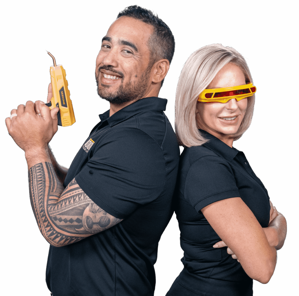 Two adults back-to-back in dark shirts; man holds a yellow power tool, smiling at the camera, while the woman wears yellow safety goggles beside him.